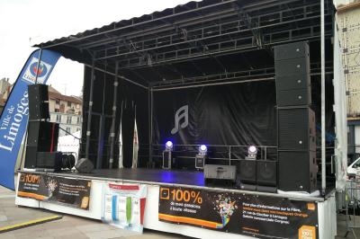 Scène extérieure vide avec équipements audio et éclairage, entourée de bannières publicitaires et d'un drapeau de la ville.