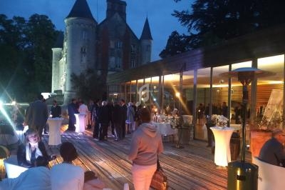 Personnes réunies en soirée sur une terrasse éclairée, avec un château en arrière-plan et des tables hautes.
