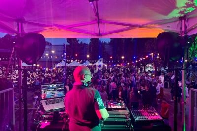Un DJ mixe sur scène lors d'un événement en plein air, avec une foule dansante et des lumières colorées.