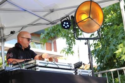 Un DJ mixe de la musique en plein air sous une tente, avec un projecteur allumé et des arbres en arrière-plan.