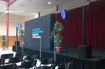 Scène avec écran affichant ’Le 9 juin, nous avons besoin d'Europe’, entourée de plantes et de chaises alignées.