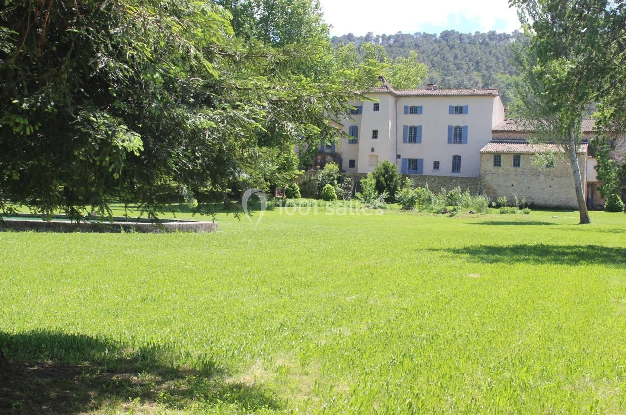 Grand jardin verdoyant avec pelouse, arbres et un bâtiment à plusieurs étages en arrière-plan sous un ciel dégagé.