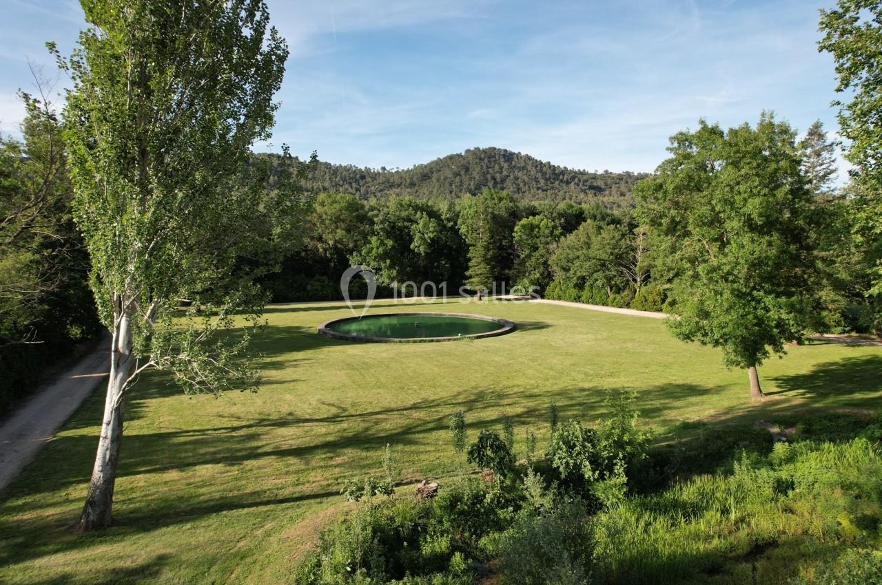 Vue d'un grand espace vert avec une pelouse, un bassin circulaire au centre et des arbres en arrière-plan.