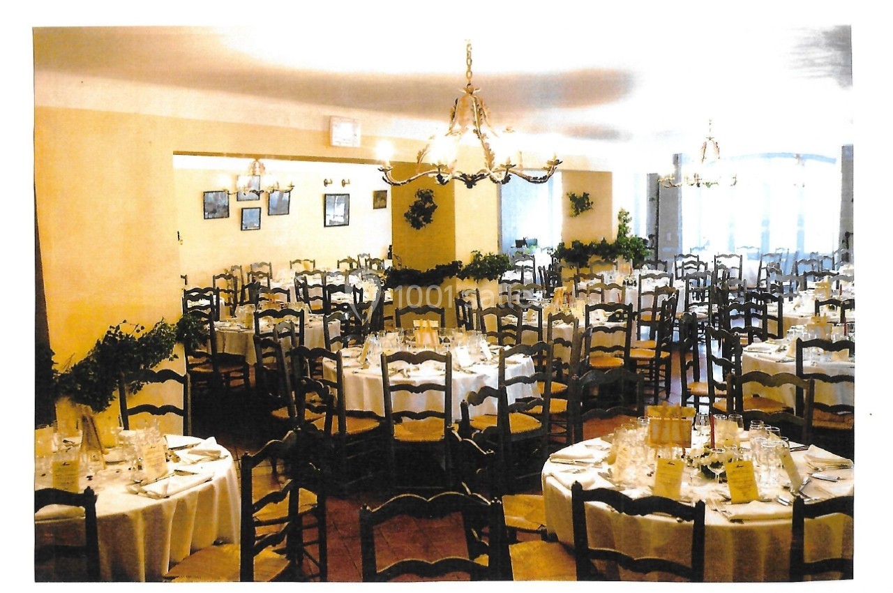 Salle de réception décorée avec des tables rondes dressées, chaises en bois et luminaires suspendus.