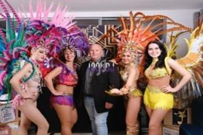 Un homme entouré de danseuses en costumes colorés avec des plumes, posant dans un décor intérieur festif.
