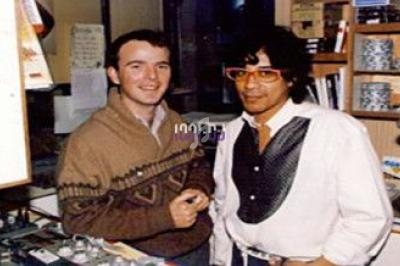 Deux hommes posent dans un studio avec des équipements audio et des étagères en arrière-plan.