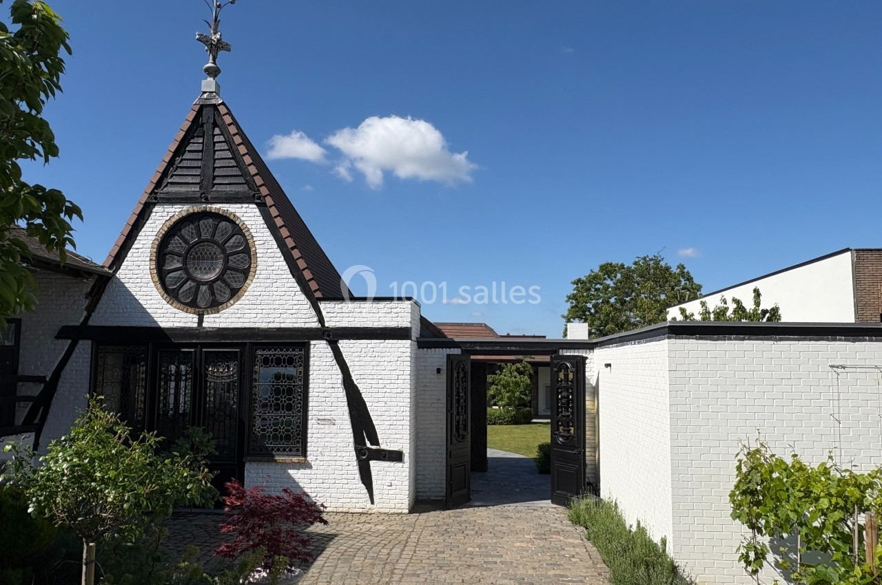 Façade d'une maison blanche avec une petite tour en briques, une rosace vitrée et un jardin pavé à l'avant.
