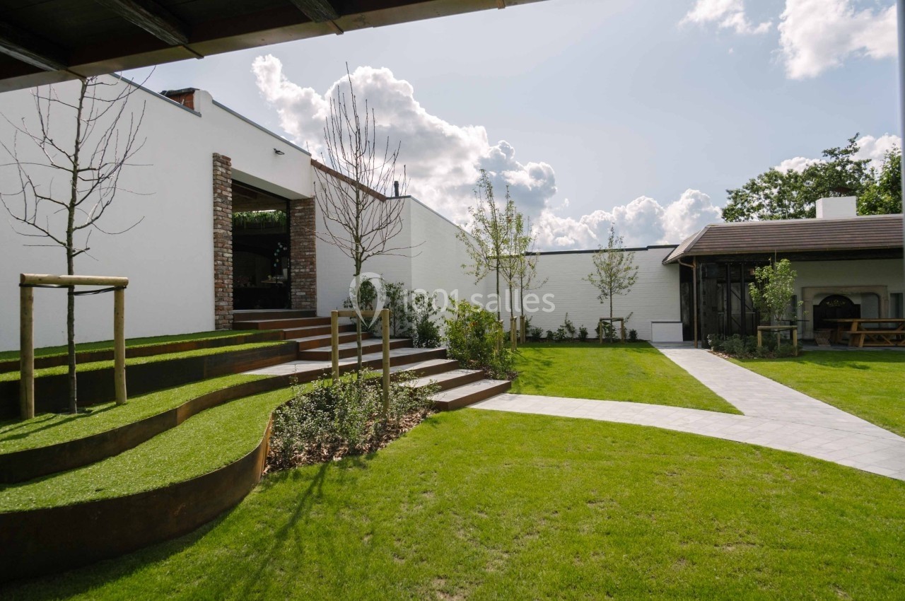 Jardin avec pelouse, allées pavées, terrasses en bois et arbres jeunes, entouré de murs blancs et bâtiments modernes.