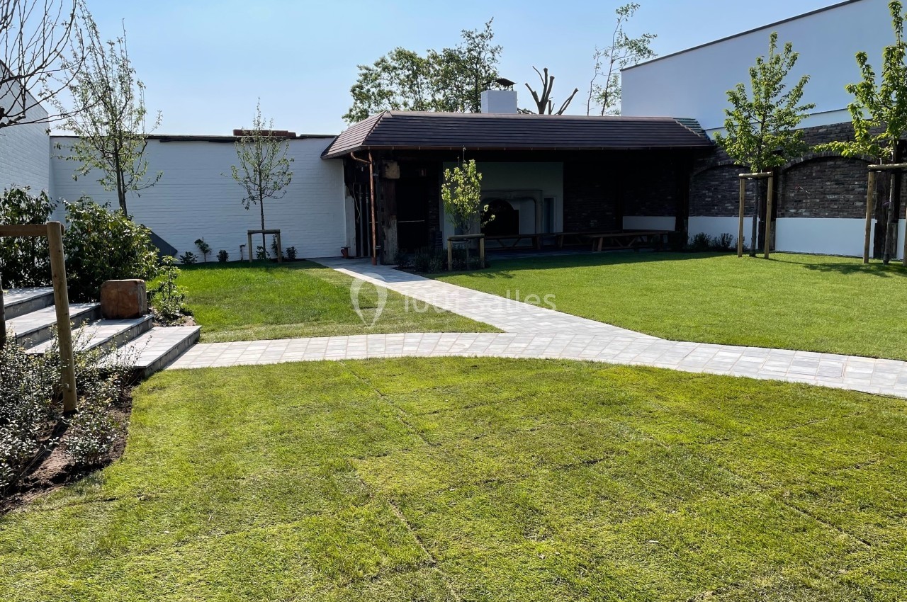 Jardin avec pelouse fraîchement posée, allées pavées et petite structure en bois adossée à un mur blanc.