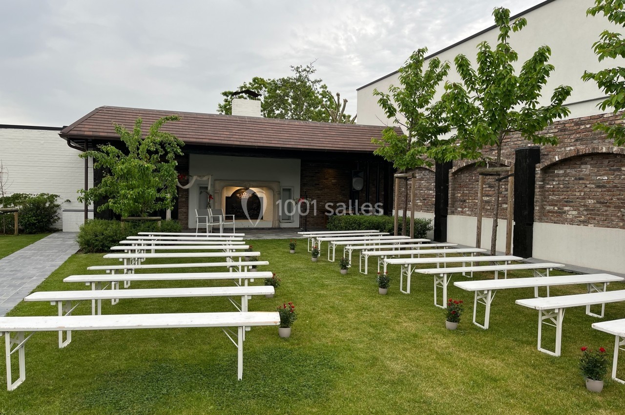 Bancs blancs disposés en rangées sur une pelouse, face à une scène couverte en bois dans un jardin extérieur.
