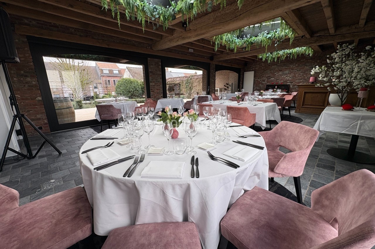 Salle de réception avec table ronde dressée, chaises roses, décoration florale et plafond orné de verdure.