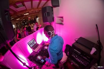 Un DJ mixe de la musique sur une console dans une salle éclairée par des lumières roses, avec des invités en arrière-plan.