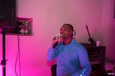 Un homme en chemise bleue chante au micro avec un casque audio, éclairé par une lumière rose dans une pièce.
