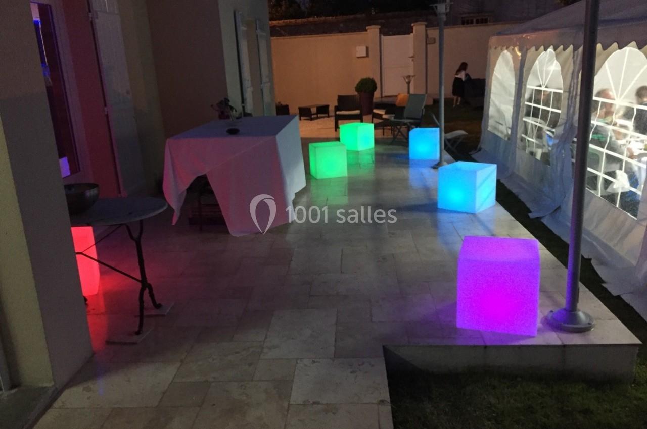 Cubes lumineux colorés disposés sur une terrasse en soirée, près d'une tente éclairée et d'une table dressée.
