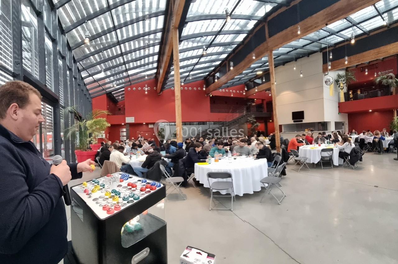 Un homme parle devant un public assis à des tables rondes dans une grande salle lumineuse avec verrière.