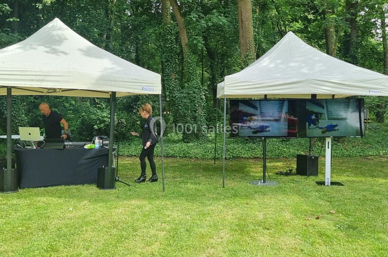 Deux tentes blanches sur une pelouse, l'une avec un stand DJ, l'autre avec un écran affichant des vidéos, en extérieur boisé.