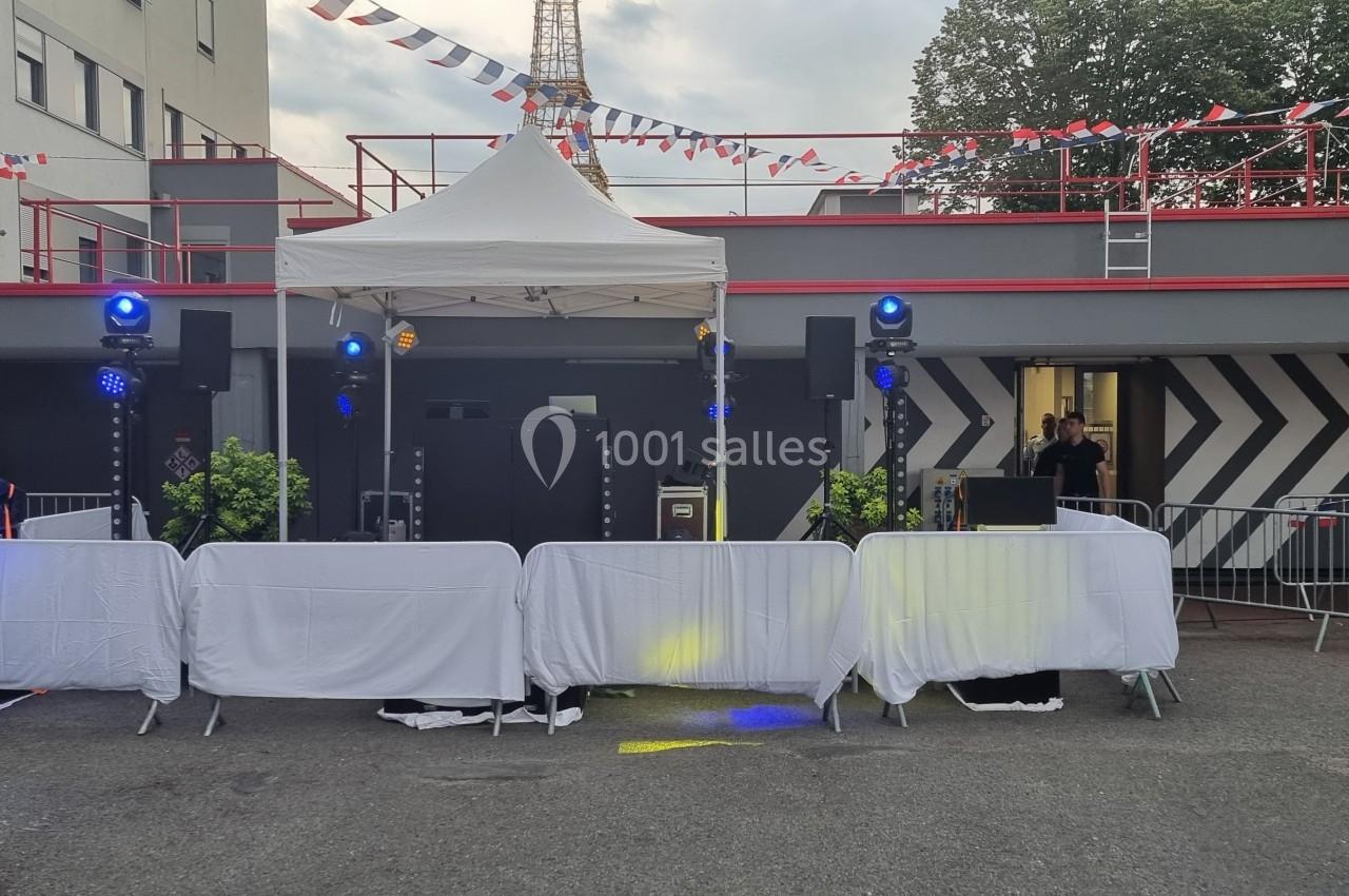 Scène extérieure avec équipement de sonorisation sous un chapiteau, décorée de guirlandes et entourée de barrières.