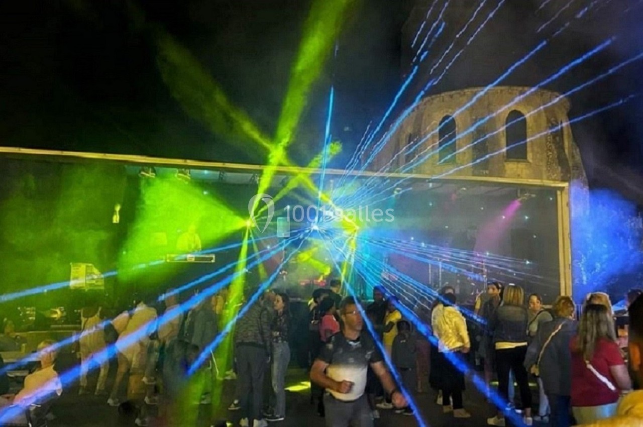 Foule rassemblée de nuit devant une scène éclairée par des faisceaux lumineux colorés, près d'un bâtiment ancien.