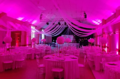 Salle de réception décorée avec des tables rondes, nappes blanches, chaises transparentes et éclairage rose.