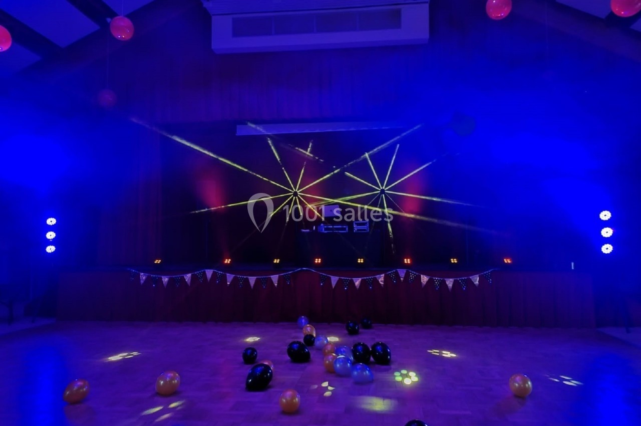 Salle de fête décorée avec des ballons au sol, éclairée par des lumières colorées et des lasers sur une scène.