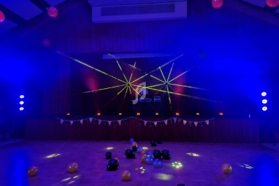 Salle de fête avec piste de danse éclairée par des lasers colorés, ballons au sol et guirlande décorative.