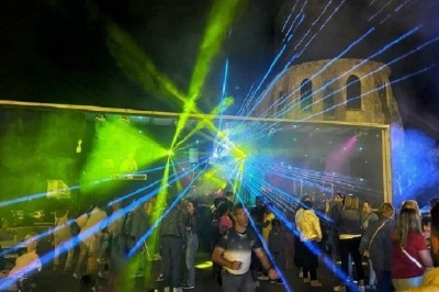 Groupe de personnes rassemblées la nuit devant une scène avec des jeux de lumière colorés et un bâtiment en arrière-plan.
