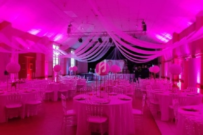 Salle de réception décorée de nappes blanches, centres de table fleuris et éclairage rose vif.
