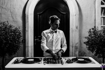 Un DJ en chemise et nœud papillon mixe sur une table de platines devant une porte voûtée.