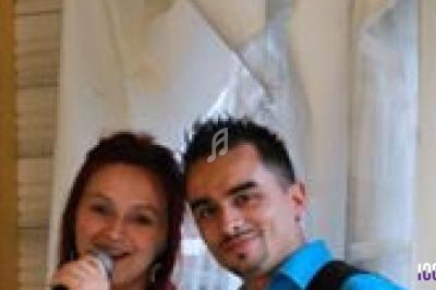 Un homme et une femme souriants, la femme tenant un micro, devant un fond clair avec des rideaux.