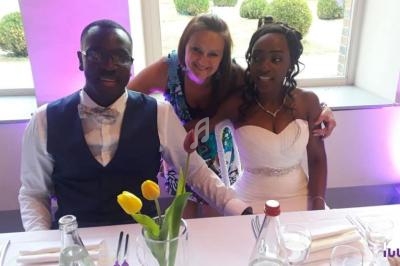 Un couple assis à une table de réception avec une femme debout derrière eux, décorée de fleurs et éclairée en violet.