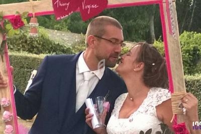 Un couple en tenue de mariage pose en souriant dans un cadre décoré avec des fleurs et des inscriptions.