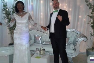 Un couple en tenue de mariage pose devant un canapé blanc décoré, avec un fond de rideaux lumineux.