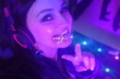 Femme portant un casque audio, souriant et faisant un signe de paix dans un environnement éclairé par des lumières colorées.