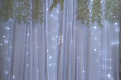 Rideau blanc décoré de guirlandes lumineuses, fleurs suspendues et initiales rouges en ruban.