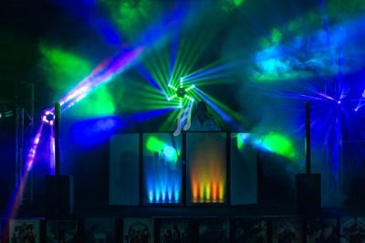 Un DJ mixe sur une scène éclairée par des faisceaux lumineux colorés dans une ambiance de soirée festive.