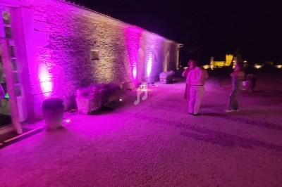 Façade en pierre éclairée par des lumières violettes, avec des personnes discutant dans une cour de nuit.