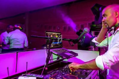 Un DJ mixe de la musique sur une console dans une salle éclairée par des lumières colorées, avec des invités en arrière-plan.