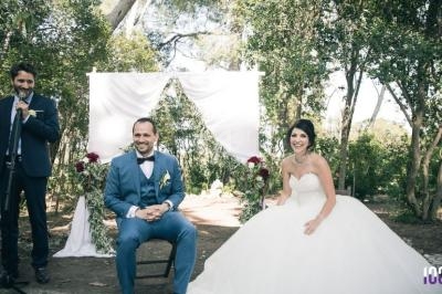 Un couple en tenue de mariage assis devant une arche décorée, avec un homme tenant un micro à gauche.