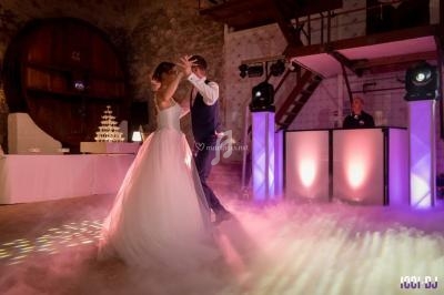 Un couple danse leur première danse de mariage dans une salle éclairée, entourée de fumée et de lumières colorées.