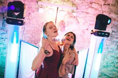 Deux femmes chantent et dansent devant un DJ dans une ambiance festive avec éclairages colorés.