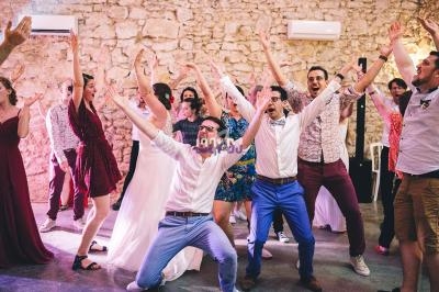 Des personnes dansent et lèvent les bras lors d'une fête dans une salle aux murs en pierre.