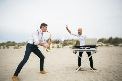 Un saxophoniste joue face à un DJ avec un ordinateur portable sur une plage sous un ciel nuageux.