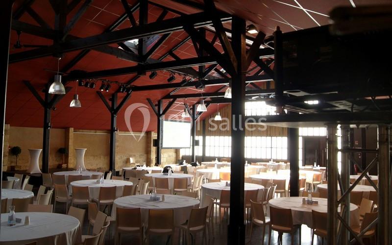 Salle de réception avec des tables rondes dressées, éclairée par des fenêtres et des lumières suspendues.