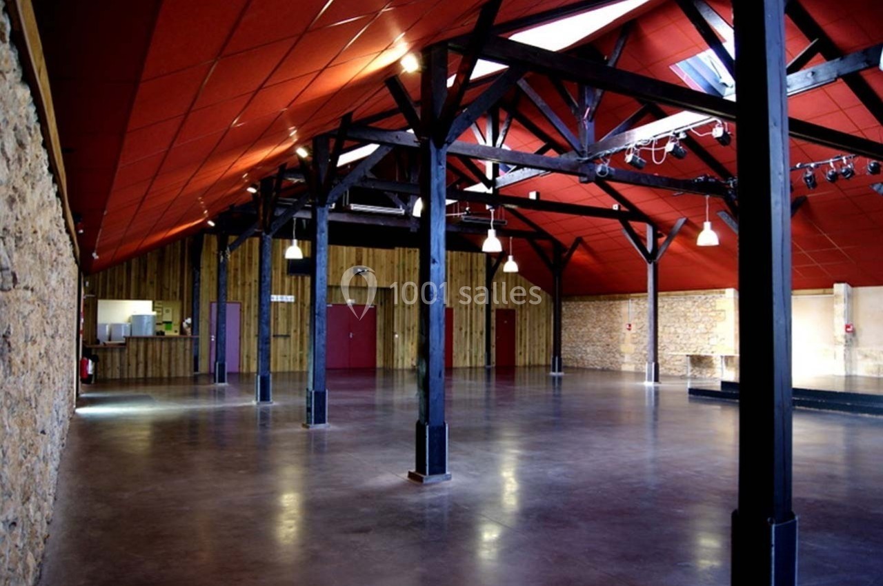 Salle spacieuse avec poutres apparentes, murs en pierre et bois, sol en béton lisse, éclairée par des luminaires suspendus.