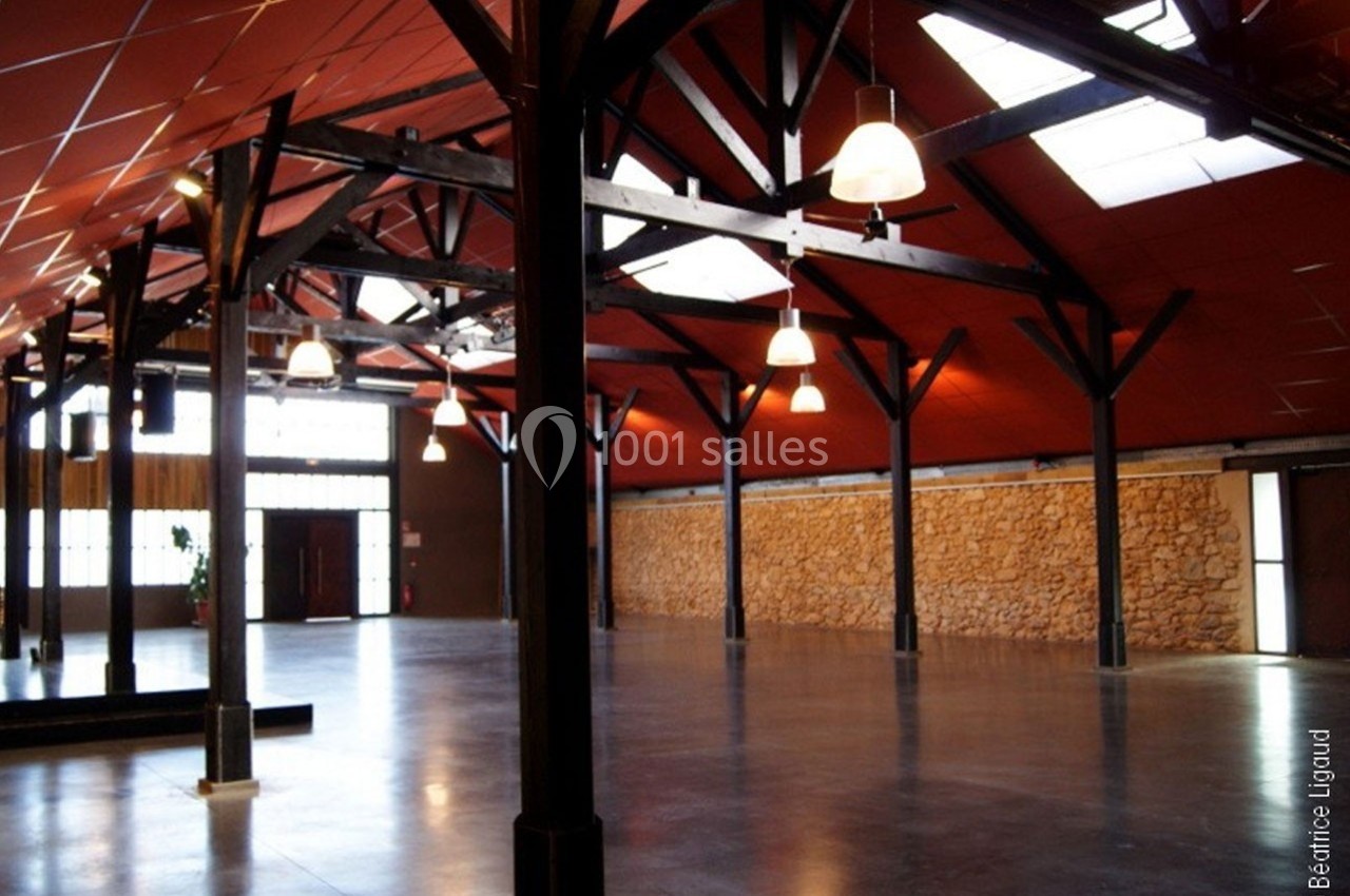 Grande salle avec charpente en bois apparente, murs en pierre et plafond rouge, éclairée par des suspensions et des fenêtres.