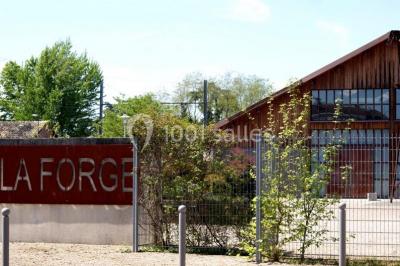 Bâtiment en bois avec une clôture métallique et un panneau indiquant ’La Forge’, entouré de végétation.