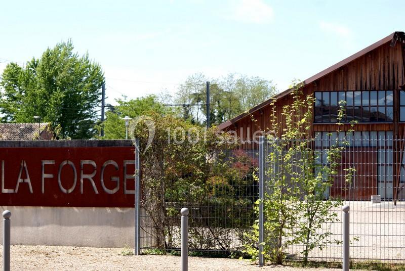 Bâtiment en bois avec une clôture métallique et un panneau indiquant ’La Forge’, entouré de végétation.