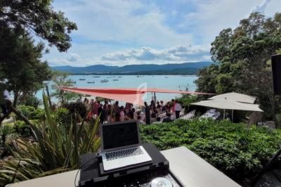 Vue d'une réception en plein air près de la mer, avec des invités rassemblés sous des voiles d'ombrage et un équipement DJ…