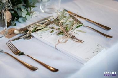 Menu décoré de branches d'olivier posé sur une table dressée avec couverts en métal et nappe blanche.