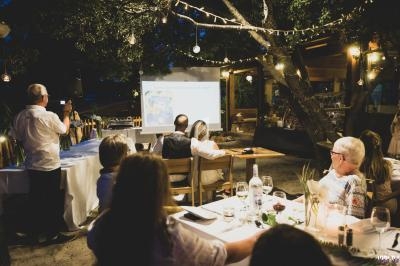 Un groupe de personnes assises à l'extérieur, regardant une présentation projetée sur un écran en soirée.
